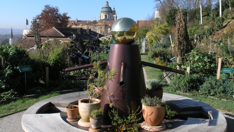 Paradiesgarten, © Martin Rotheneder Ein dekorativer Garten mit einer Skulptur im Vordergrund und einem historischen Gebäude im Hintergrund.