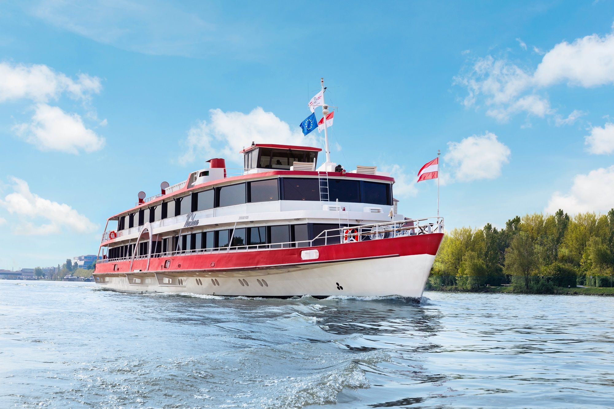 Ein Passagierschiff mit roten und weißen Farben fährt auf einem Fluss unter blauem Himmel.