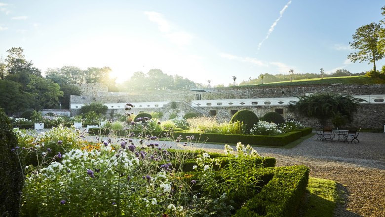 Ein bl&uuml;hender Garten mit gepflegten Hecken und Blumenbeeten vor einer historischen Mauer bei Sonnenaufgang.