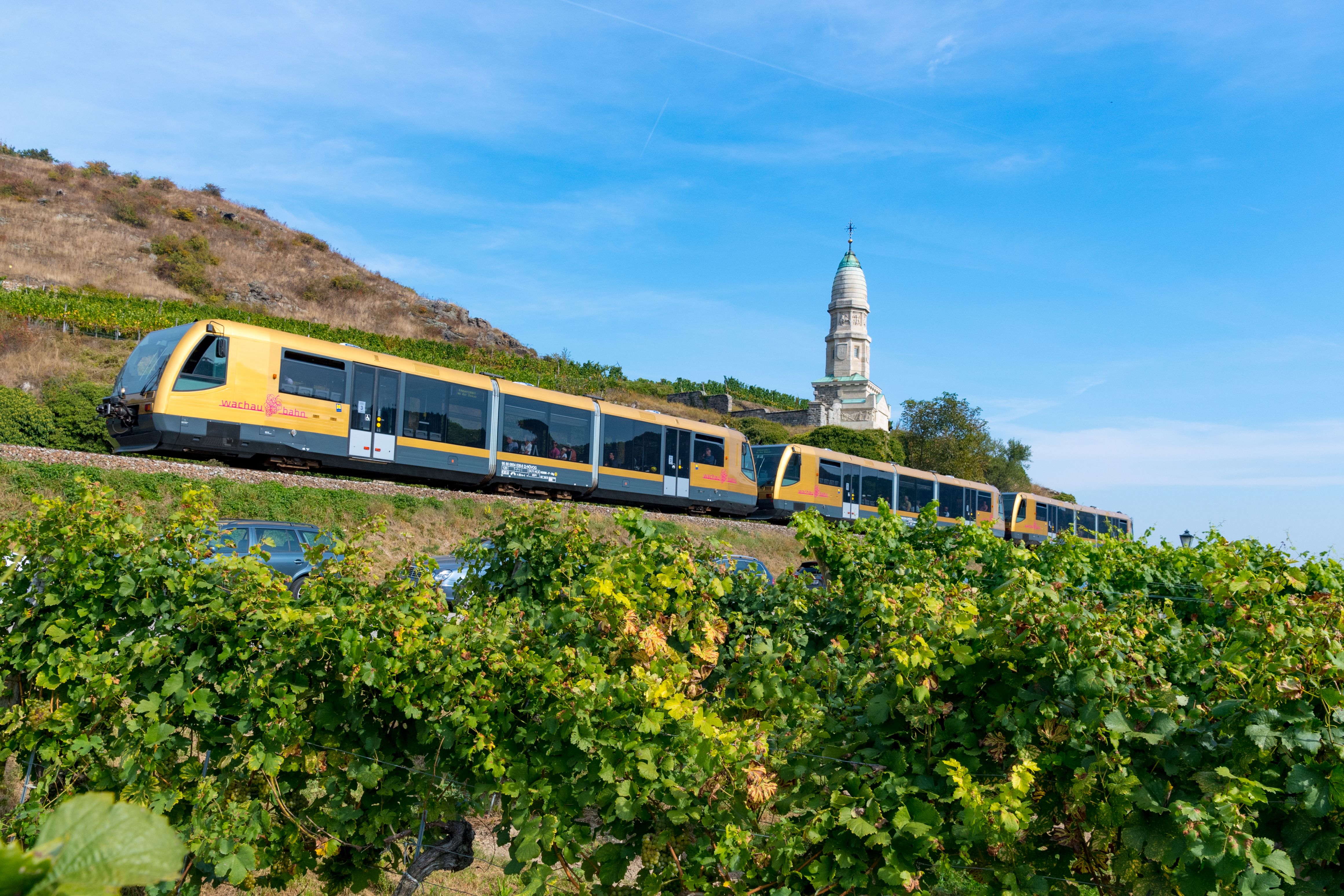 Ein gelb-grauer Zug der WEin goldener Zug der Wachaubahn fährt durch die Weinberge der Wachau. Im Hintergrund erhebt sich das Franzosendenkmal.achaubahn fährt durch die Weinberge der Wachau. Im Hintergrund erhebt sich eine markante weiße Kirche auf einem Hügel unter blauem Himmel.