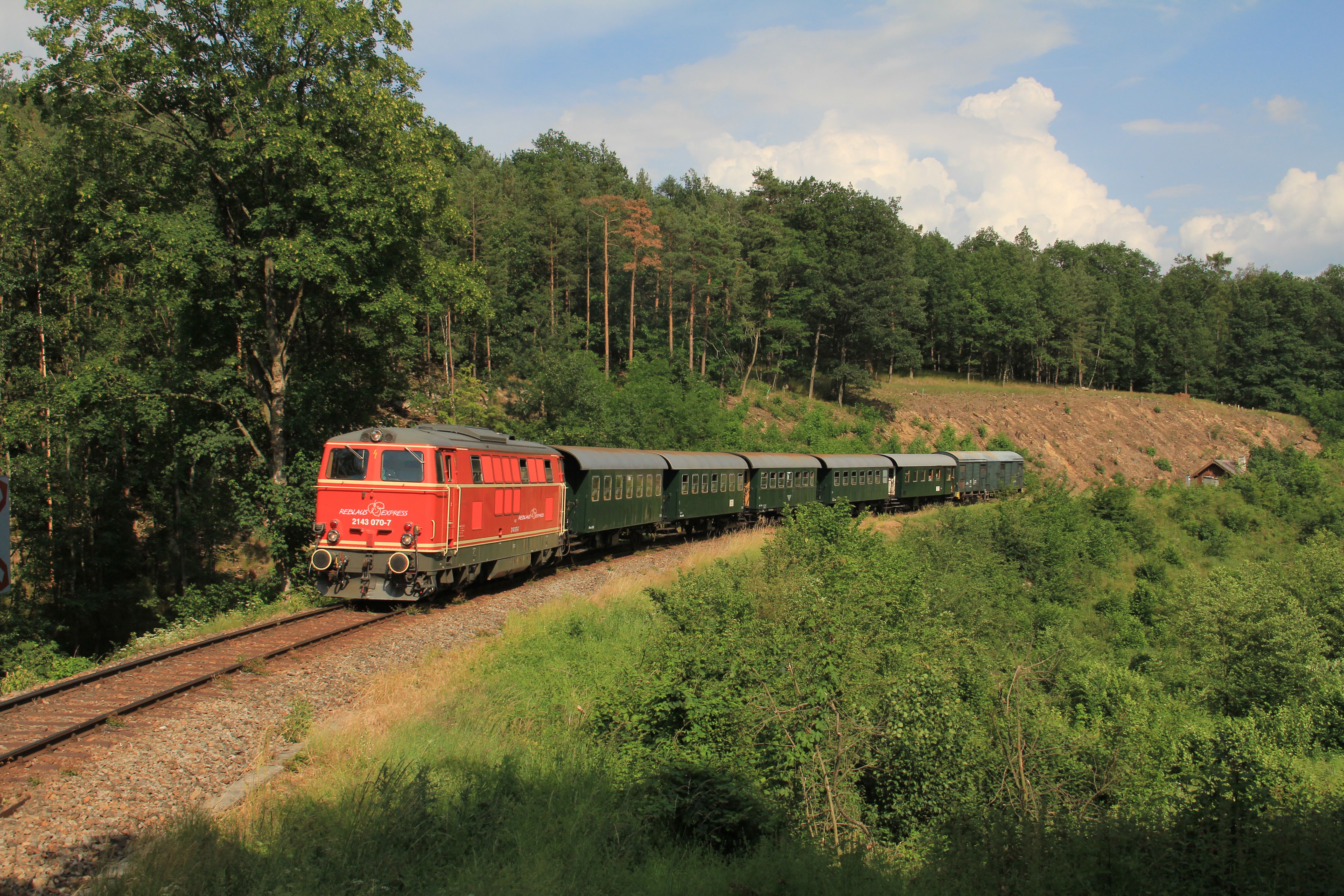 Eine rote Diesellokomotive des Reblaus Express zieht mehrere grüne Personenwagen auf einer eingleisigen Bahnstrecke durch eine bewaldete Landschaft.