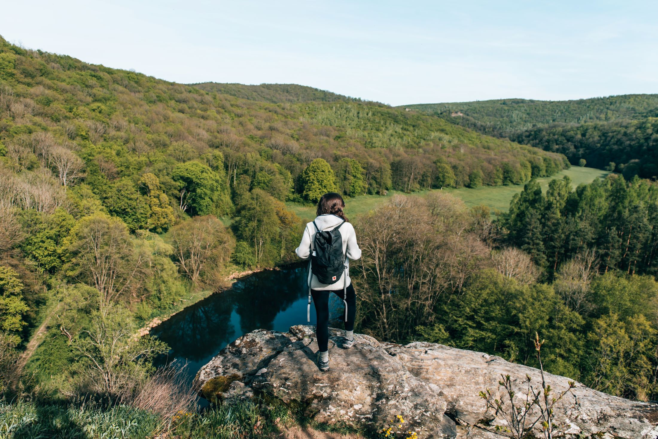 Person mit Rucksack steht auf einem Felsen und blickt auf bewaldete Landschaft im Nationalpark Thayatal.