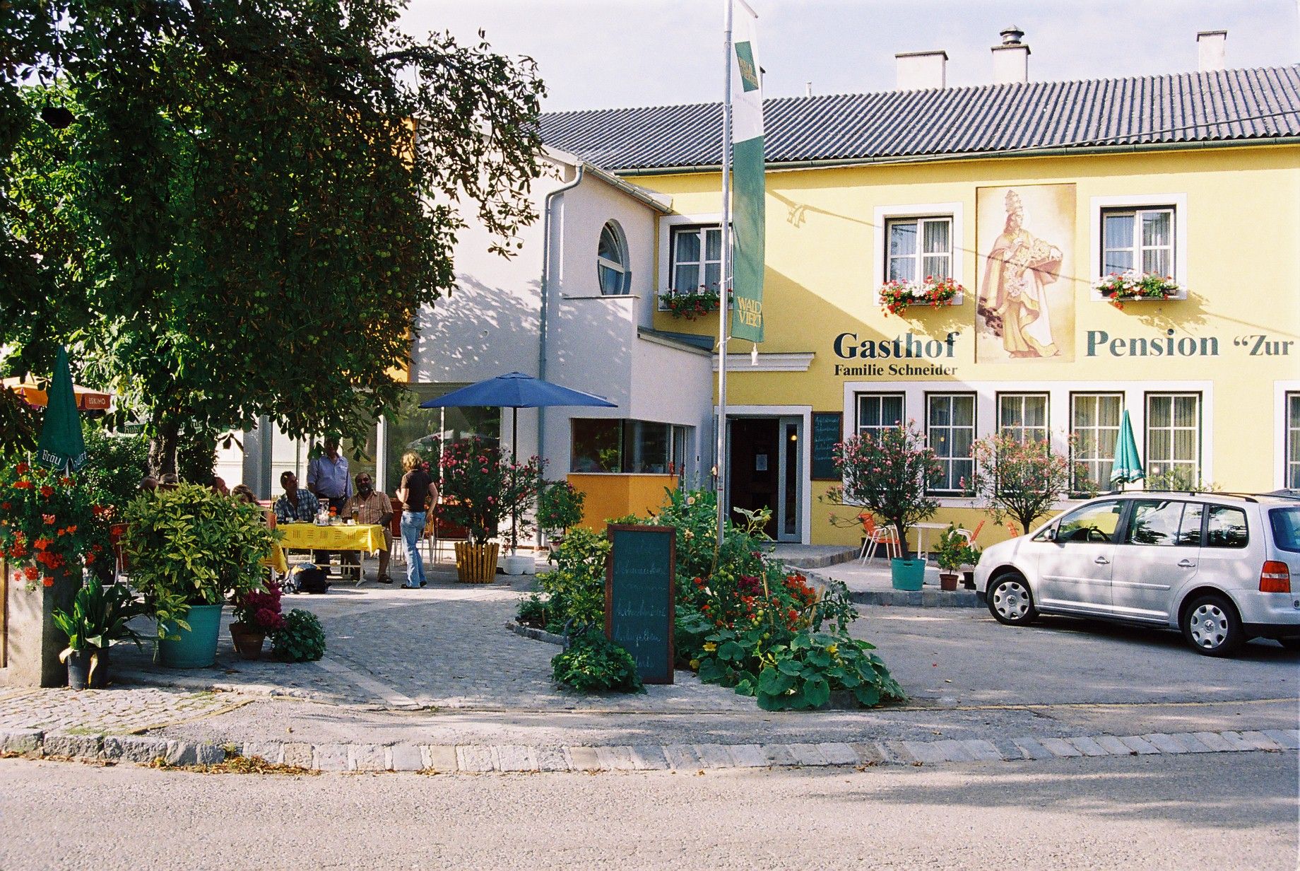 Ein gelbes Gebäude mit der Aufschrift 'Gasthof-Pension Zur Hammerschmiede'. Vor dem Gebäude stehen Tische, Stühle und ein Auto.