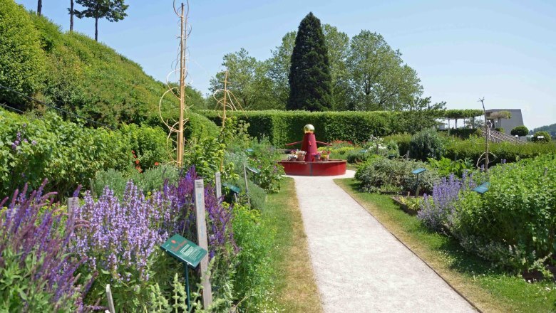 Ein gepflegter Gartenweg im Stift Melk, umgeben von blühenden Pflanzen und einem roten Brunnen im Hintergrund.