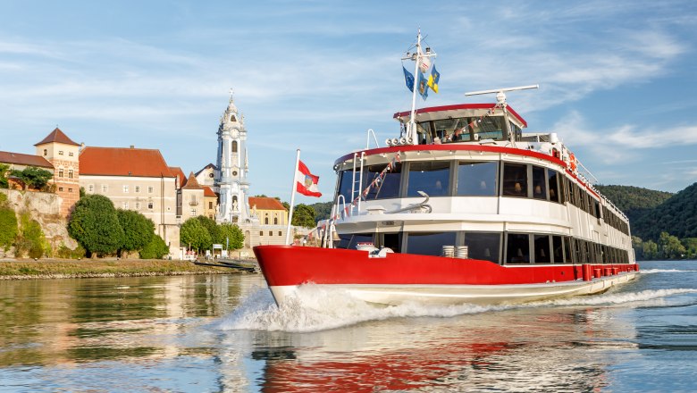 Ein rotes Passagierschiff f&auml;hrt auf der Donau vor einer historischen Stadt mit einem markanten Kirchturm.