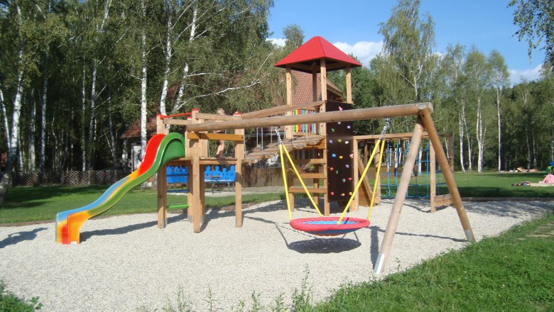 Ein Kinderspielplatz mit Rutsche, Schaukel und Kletterger&uuml;st, umgeben von B&auml;umen.