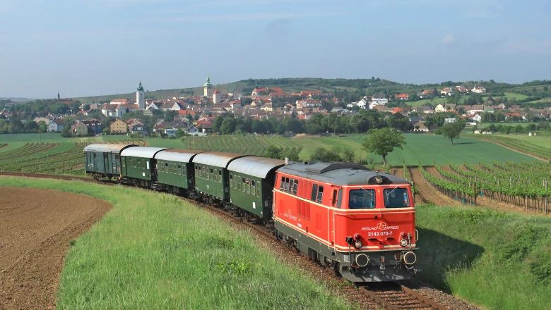 Roter Reblaus Express mit grünen Waggons fährt durch Weingärten, im Hintergrund liegt eine Ortschaft.