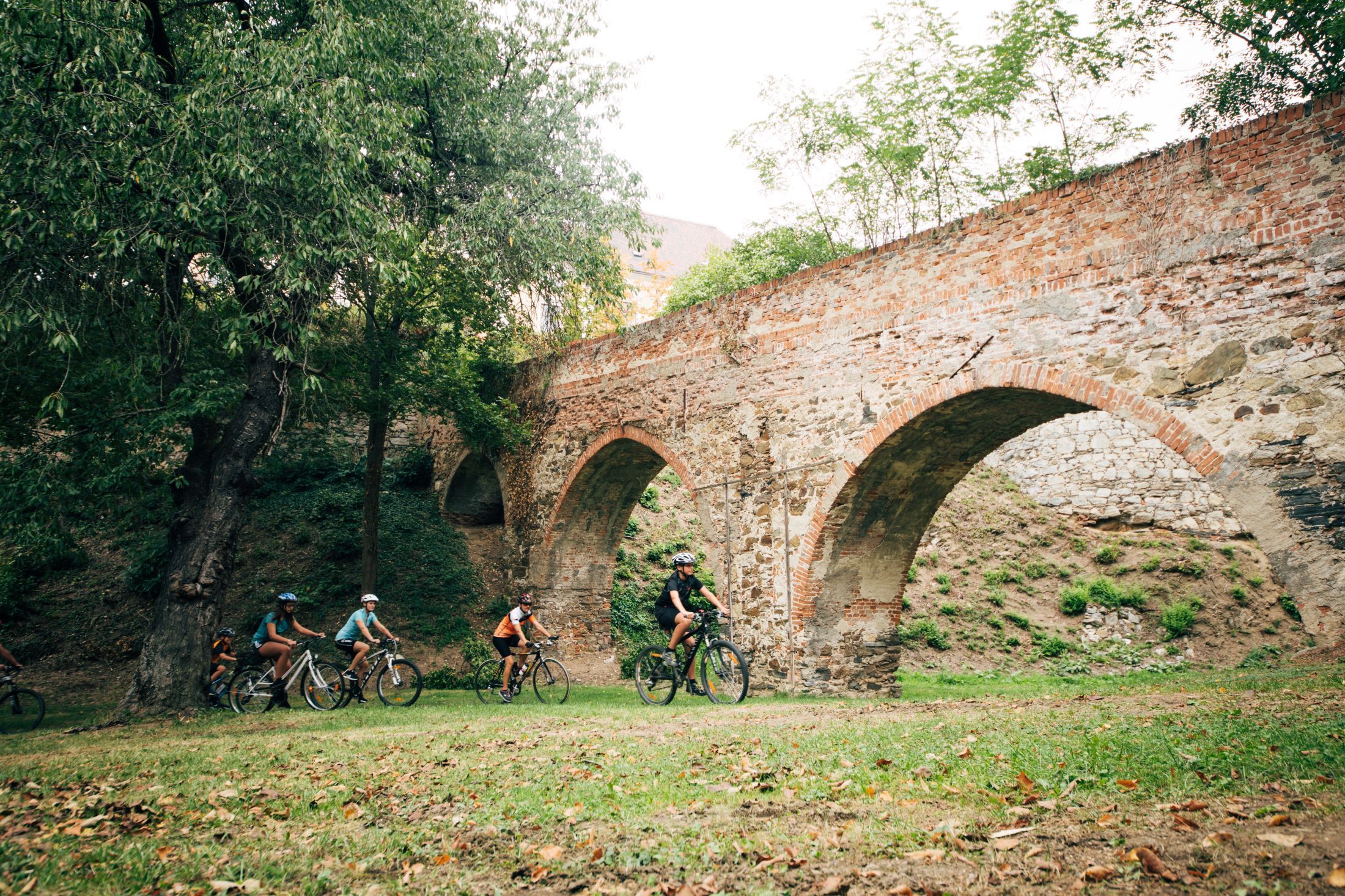 Gruppe von Radfahrern unter einer alten Backsteinbrücke im Grünen.