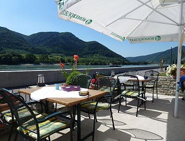 Ein Gastgarten mit Tischen und St&uuml;hlen unter Sonnenschirmen, mit Blick auf die Donau und bewaldete H&uuml;gel im Hintergrund.