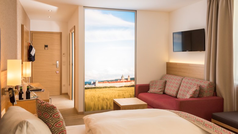 Modernes Hotelzimmer mit Doppelbett, rotem Sofa, Flachbildfernseher und gro&szlig;em Fenster mit Landschaftsausblick.