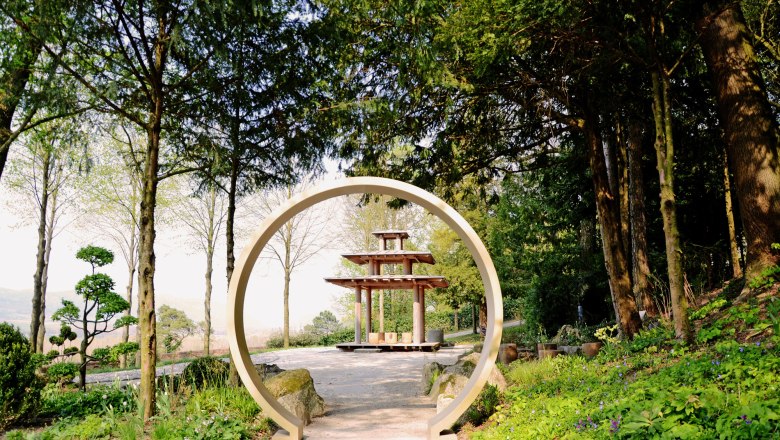 Ein orientalischer Garten mit einem runden Torbogen und einer Pagode im Hintergrund, umgeben von Bäumen und Pflanzen.