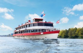 Ein Passagierschiff mit roten und wei&szlig;en Farben f&auml;hrt auf einem Fluss unter blauem Himmel.